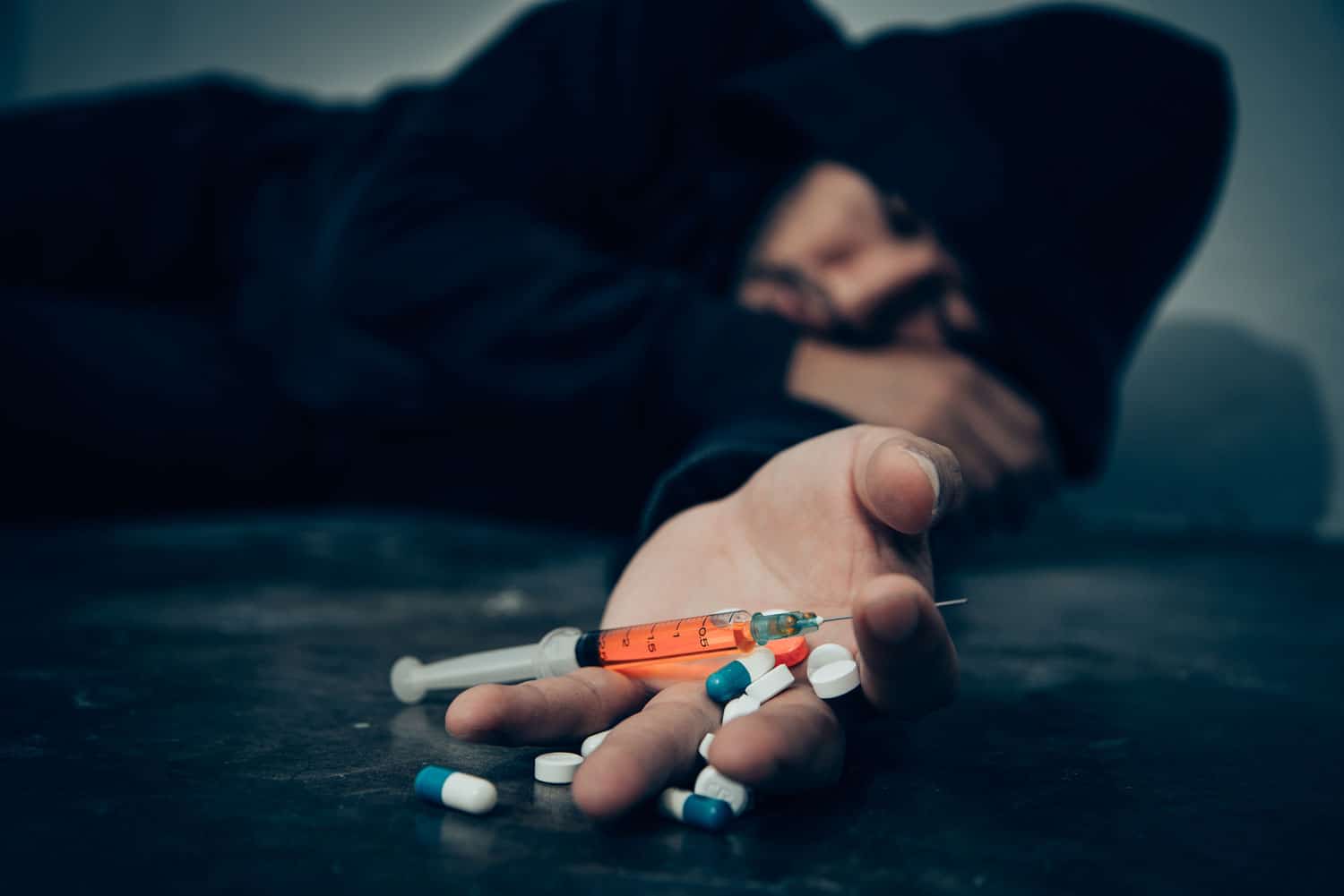 Man lying on the floor with a syringe and some drug pills in his hand.